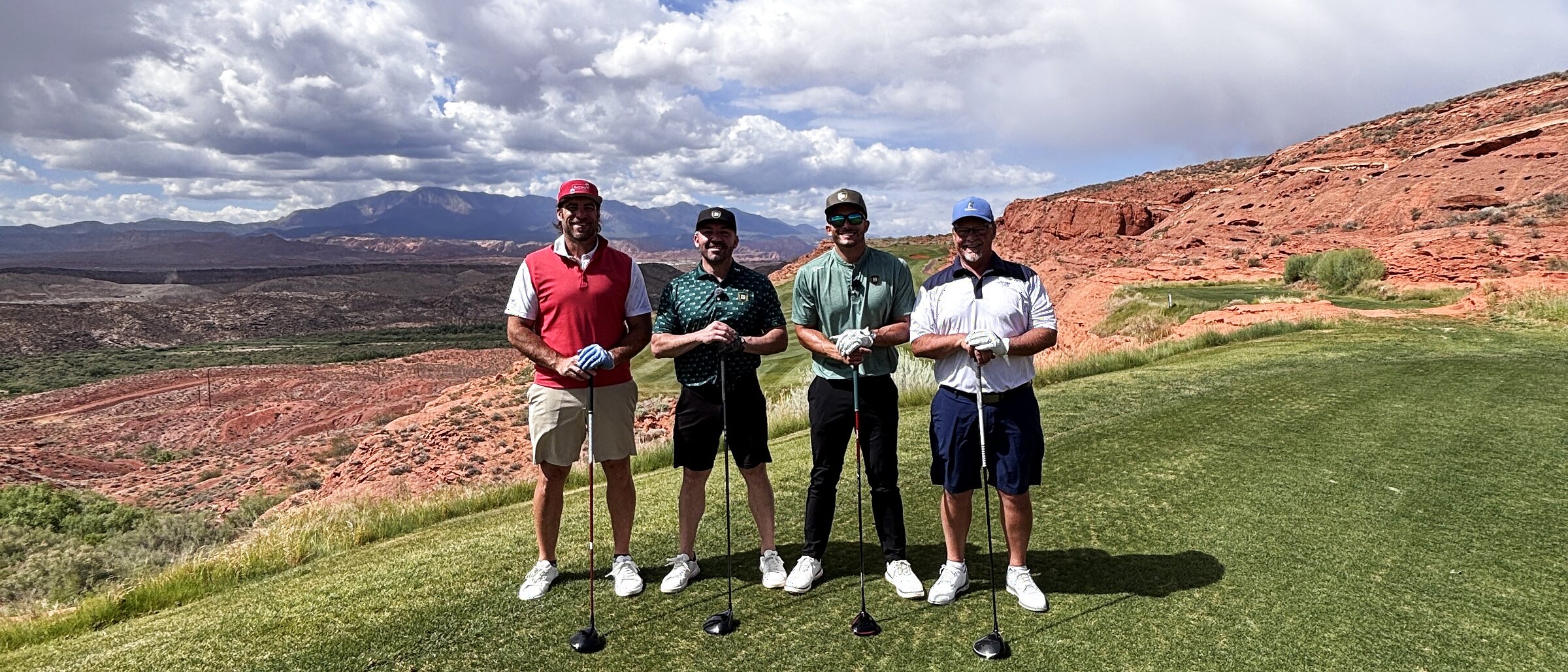A foursome standing on an elevated tee box, red sandstone cliffs and desert mountains stretching to the horizon behind them